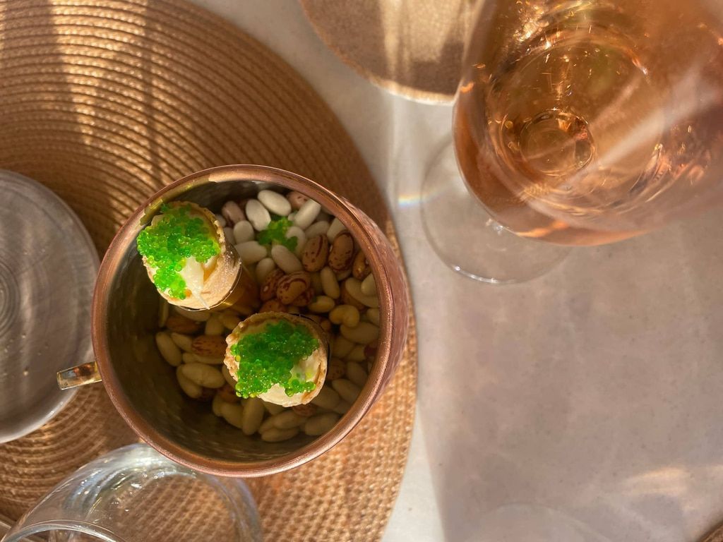 Cuenco de cobre con bocadillos, incluidos dos artículos redondos cubiertos con guarnición verde, junto con un vaso de bebida rosa.