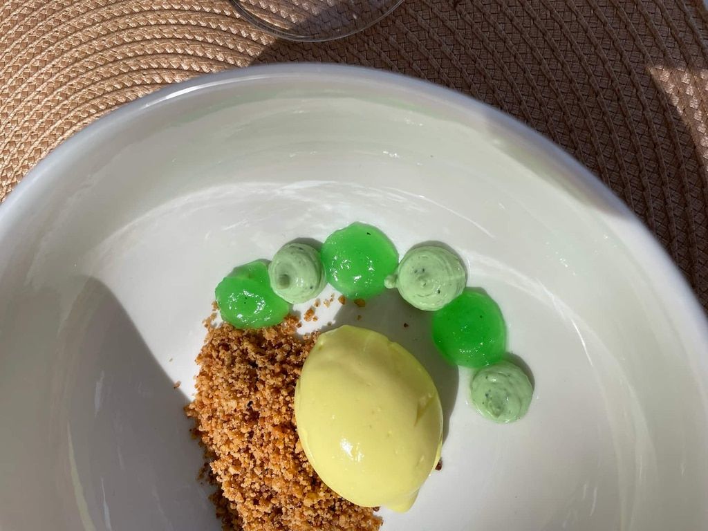 Un postre en un recipiente blanco que incluye una bola de helado amarillo, burbujas verdes tipo gel y una cobertura de migas marrones.