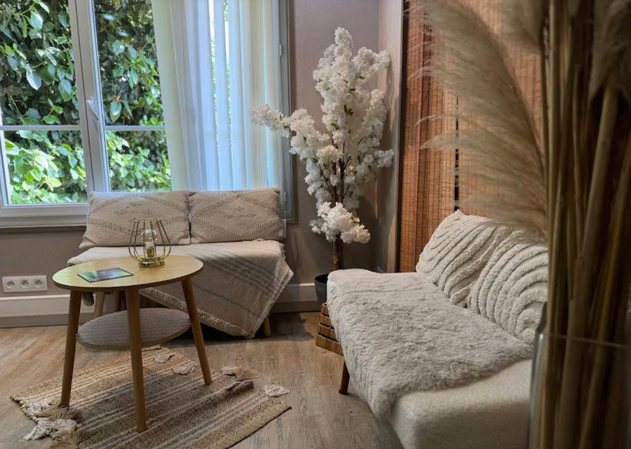Salon confortable avec fauteuils en osier, petite table ronde et grandes fenêtres avec voilages.