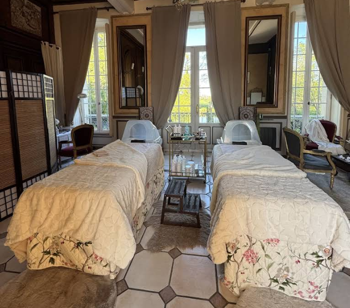 Deux tables de massage confortables dans une salle de spa baignée de lumière naturelle, avec miroirs, fenêtres et voilages.