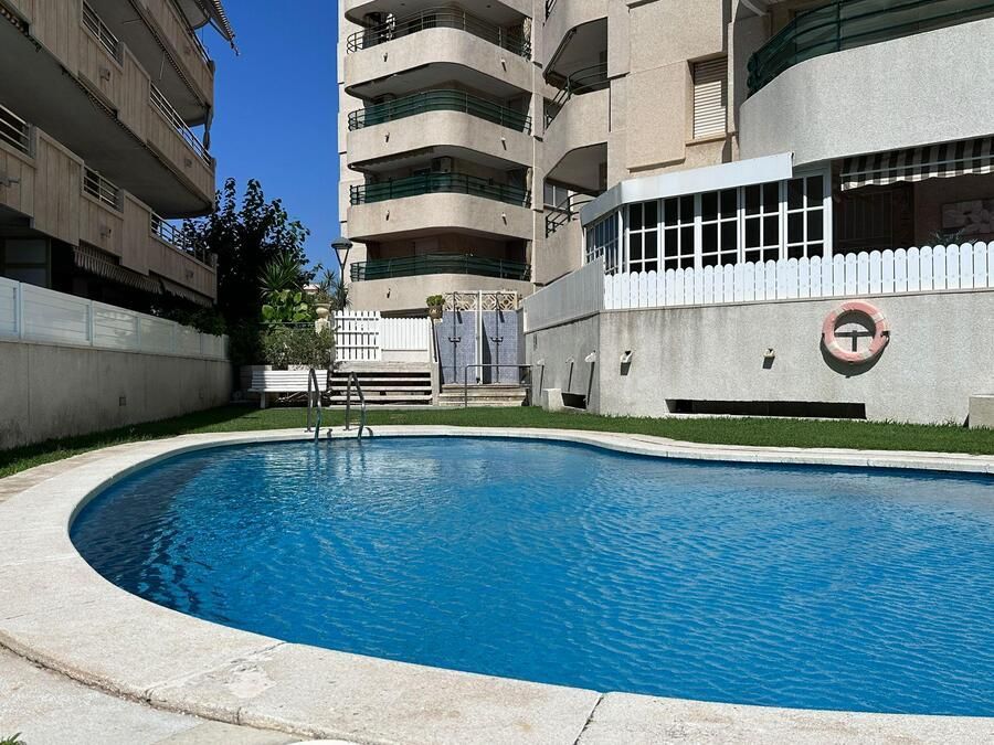Piscina frente a edificios de apartamentos. Agua azul cristalina, edificios blancos y grises, día soleado.