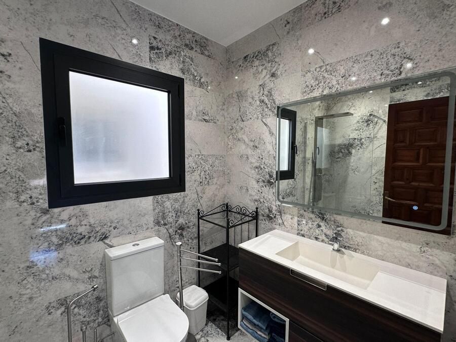 Baño con paredes con estampado de mármol, tocador oscuro, ventana cuadrada, inodoro y espejo.