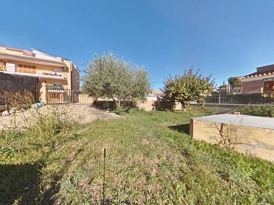 Lote cubierto de hierba con dos árboles, rodeado de edificios y un cielo azul claro.