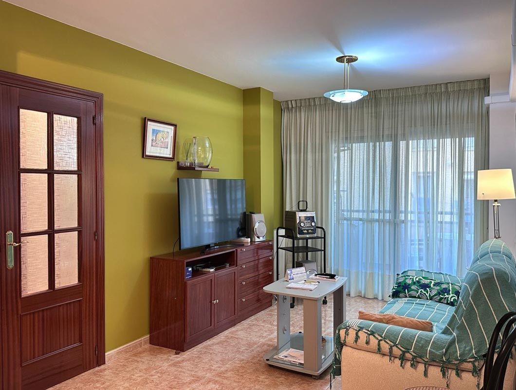 Sala de estar con paredes verdes, puerta de madera, TV, sofá y ventana con cortinas.