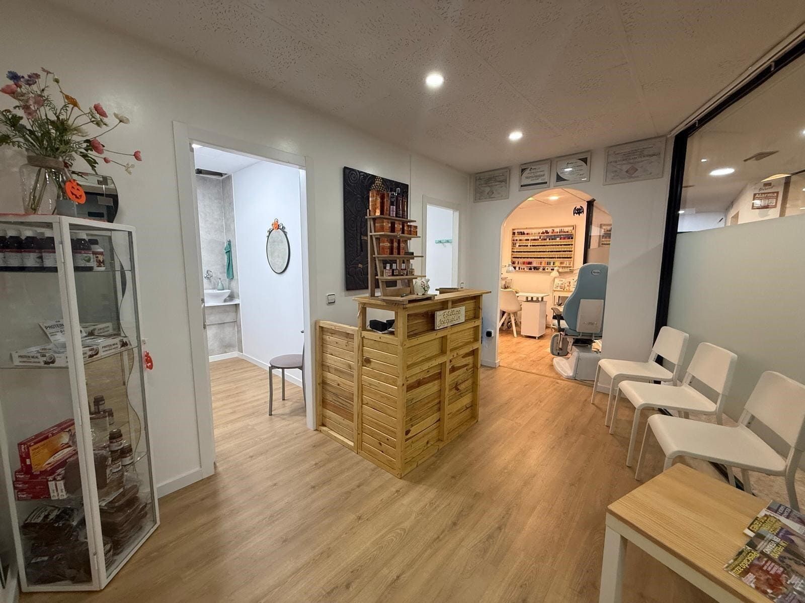 Interior de salón de uñas con pisos de madera, área de espera con sillas, mostrador de recepción y puerta arqueada al área de servicio.