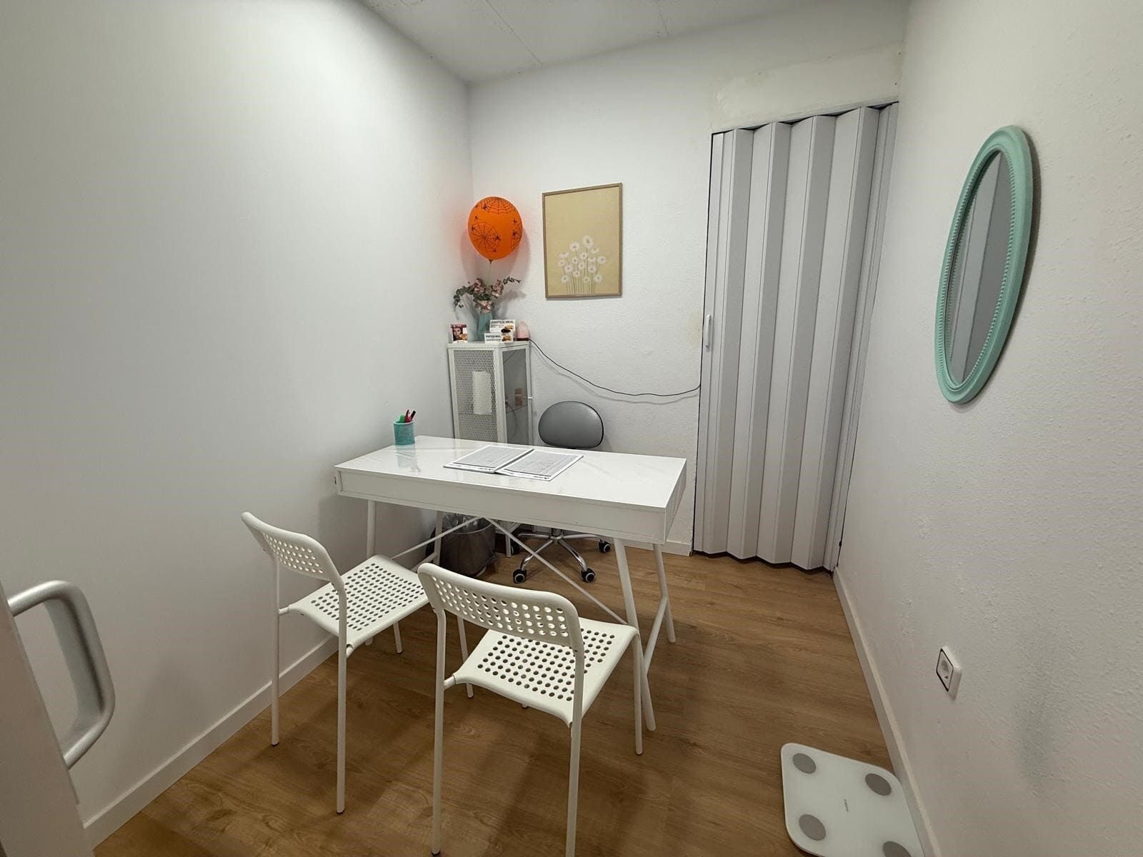 Pequeña oficina de paredes blancas con mesa, sillas, cortina, espejo, báscula y decoraciones.