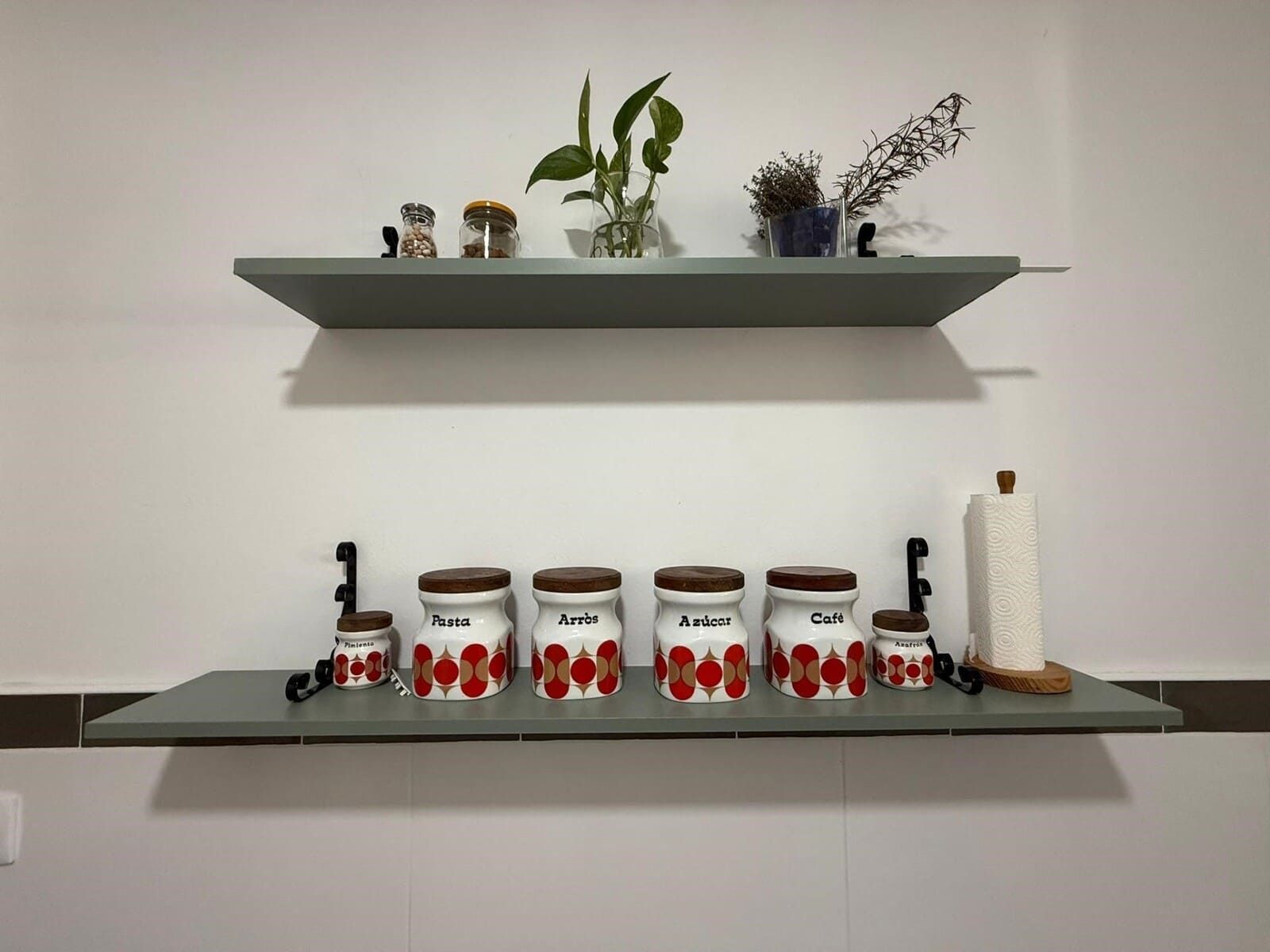Dos estantes grises sobre una pared blanca. El estante inferior contiene frascos y el superior, plantas.
