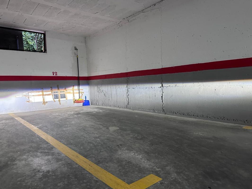 Plaza de aparcamiento interior con líneas amarillas, suelo de hormigón y paredes blancas con franja roja; escoba y ventana.