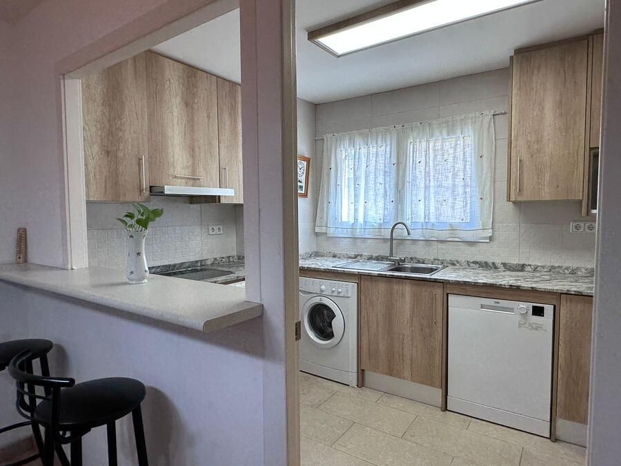 Cocina con gabinetes de madera clara, electrodomésticos, fregadero y barra de desayuno con taburetes.