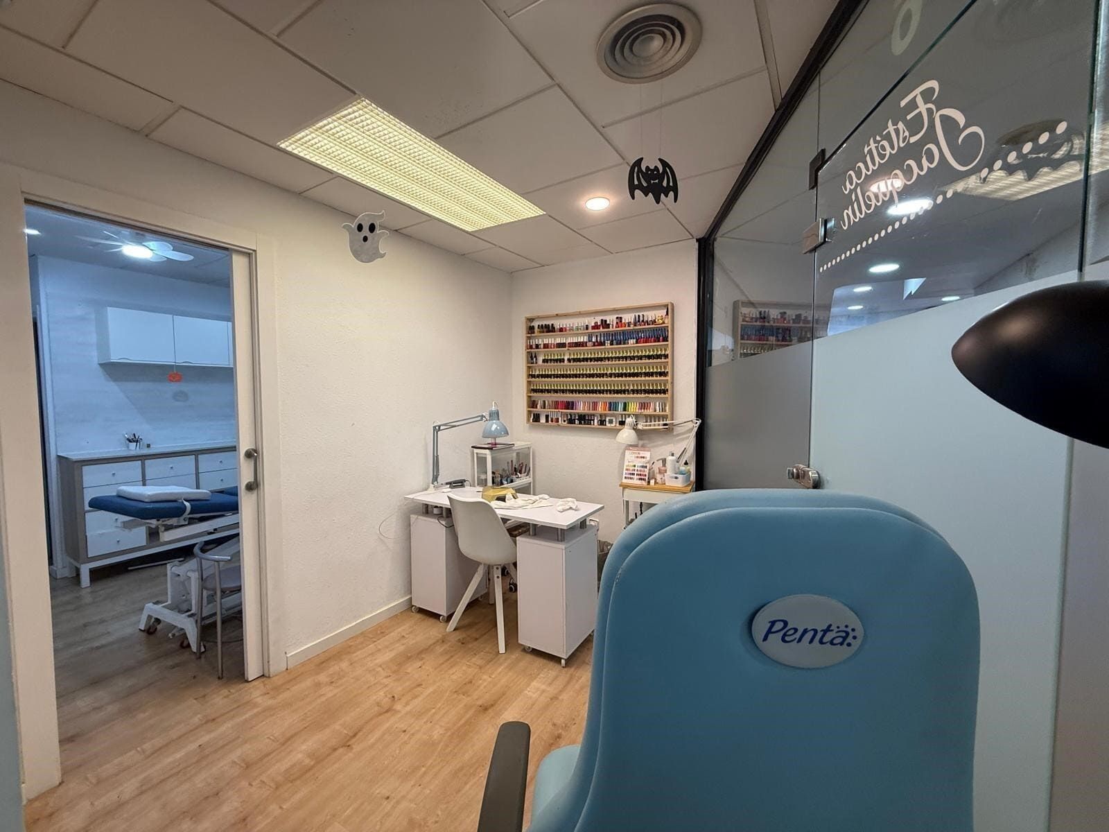 Interior de salón de uñas con una silla azul claro, un expositor de esmaltes y una estación de trabajo. Se ve otra habitación a través de una puerta.