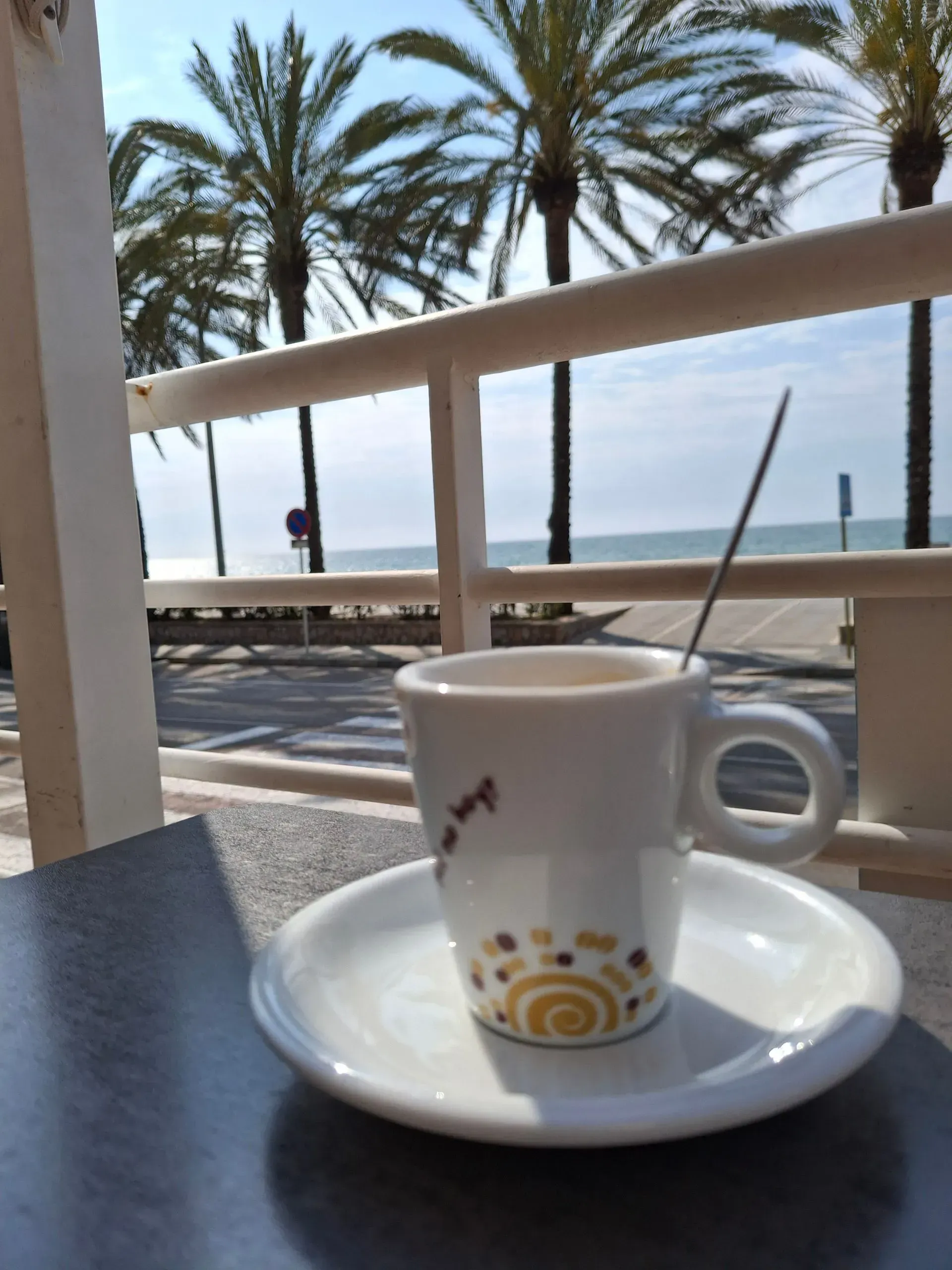 Una taza y un platillo con una cuchara sobre una mesa con palmeras al fondo.