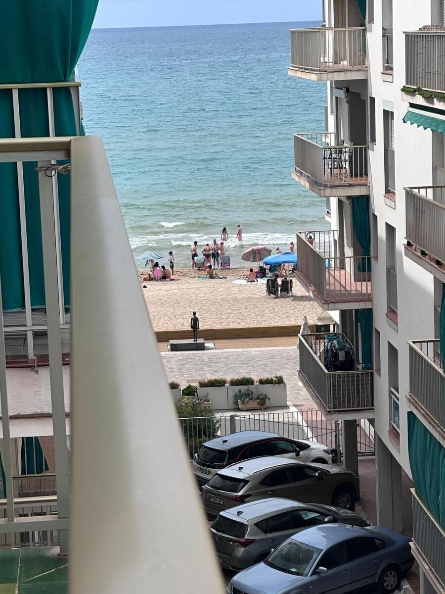 Escena de playa desde un balcón: gente en la arena, océano, edificios, coches aparcados debajo.