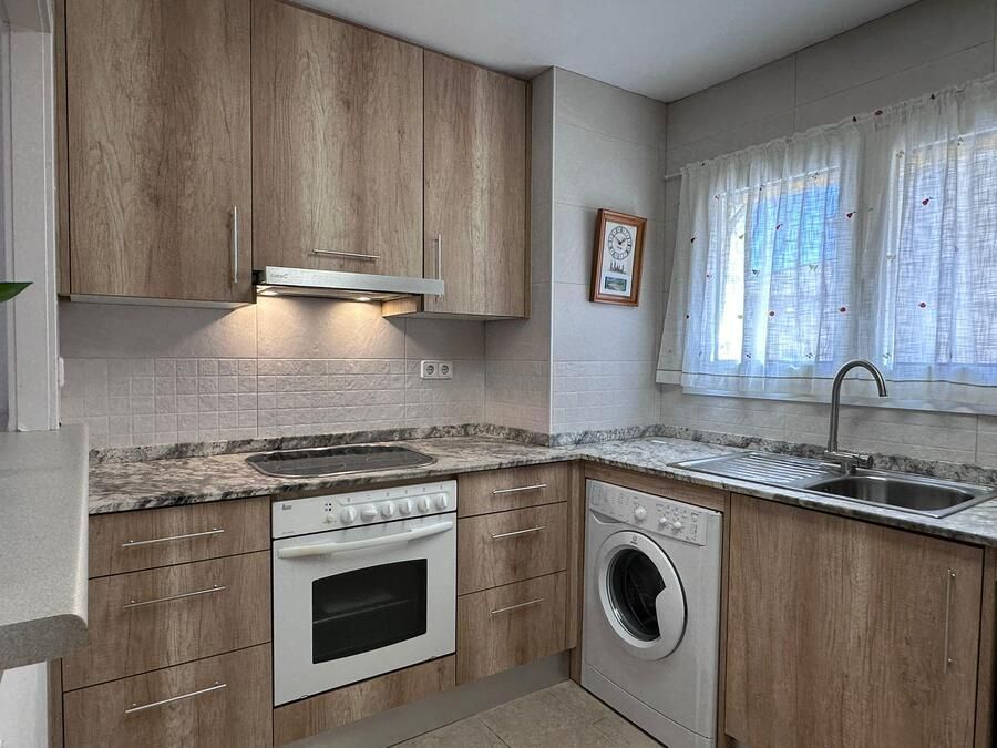 Una pequeña cocina con gabinetes de madera, una estufa y una lavadora cerca de una ventana con cortinas transparentes.