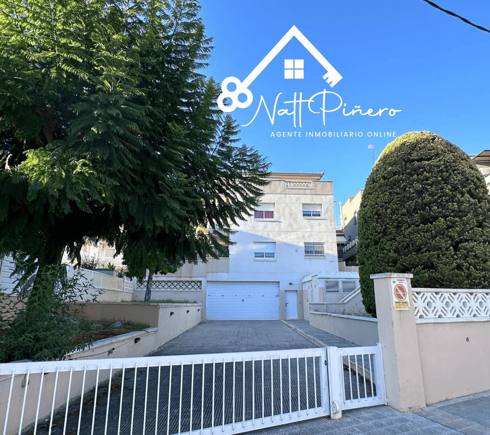 Exterior de un edificio blanco de tres pisos con entrada cerrada; árboles verdes y cielo azul.