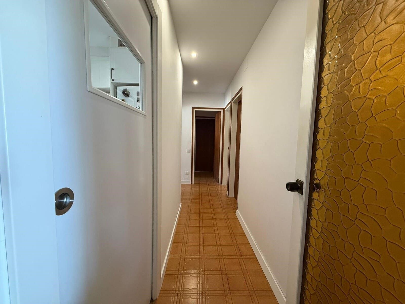 Pasillo estrecho con suelo de baldosas color canela y paredes blancas. Puertas a ambos lados, luces de techo.