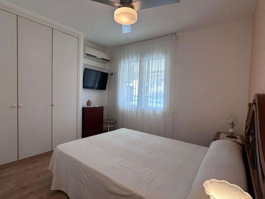 Dormitorio con cama blanca, armario, TV, ventana con cortinas transparentes y ventilador de techo.