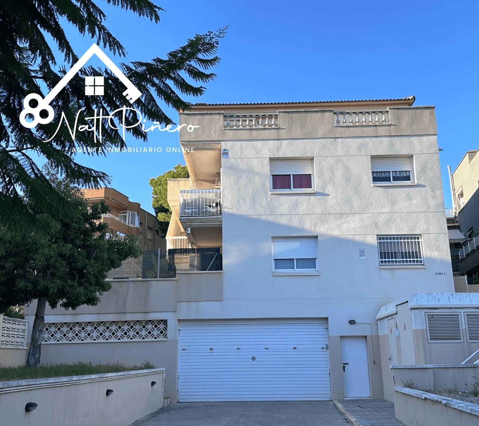 Edificio de tres pisos con garaje, balcón y logo frente a un cielo azul.