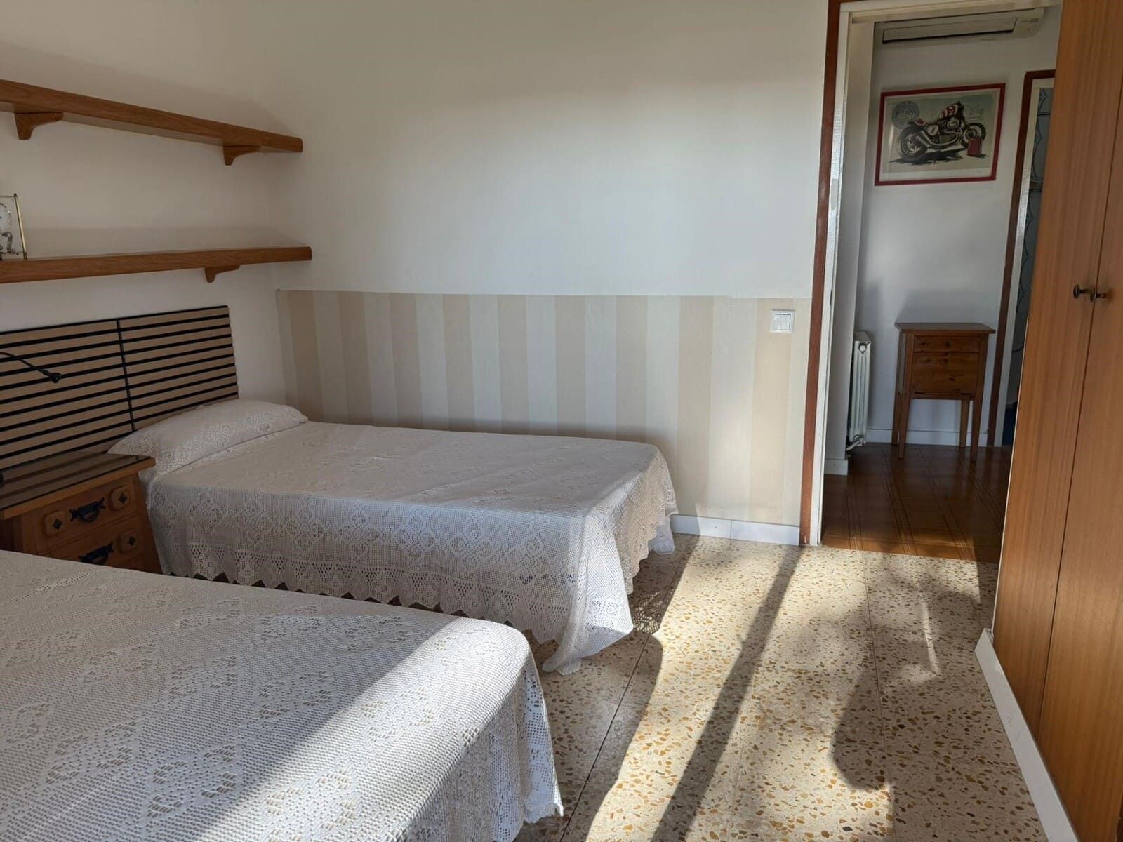 Camas individuales en un dormitorio con estantes de madera, paredes neutrales y una puerta a un pasillo.