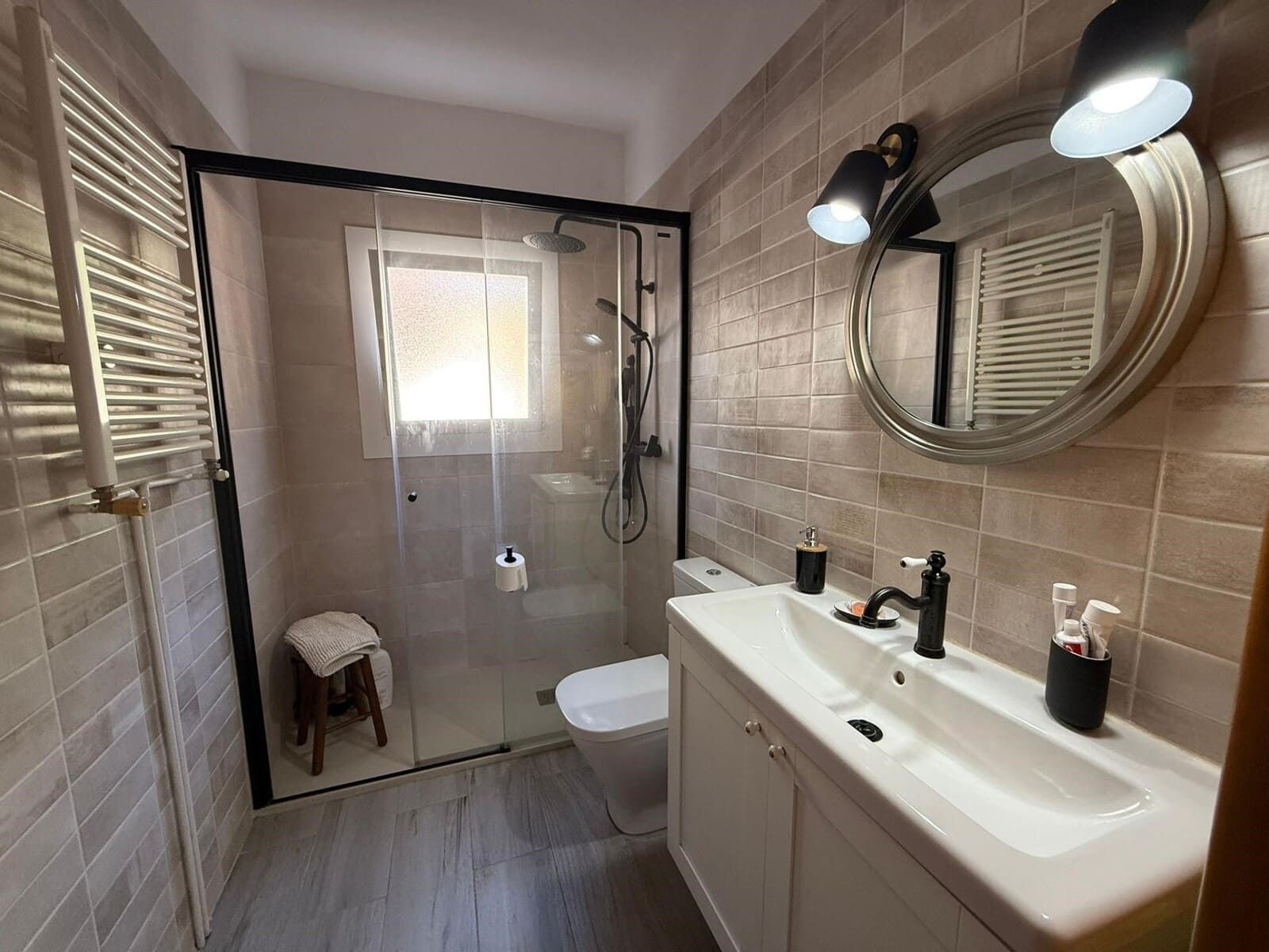 Baño con ducha, lavabo, inodoro y espejo redondo. Paredes de azulejos beige con detalles en blanco y gris.