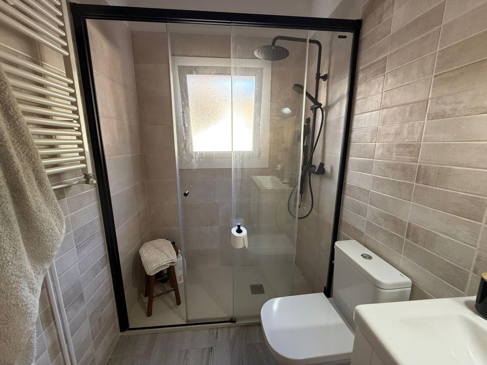 Baño con ducha de cristal, marco negro, inodoro, toallero y taburete pequeño. Paredes de azulejos beige.
