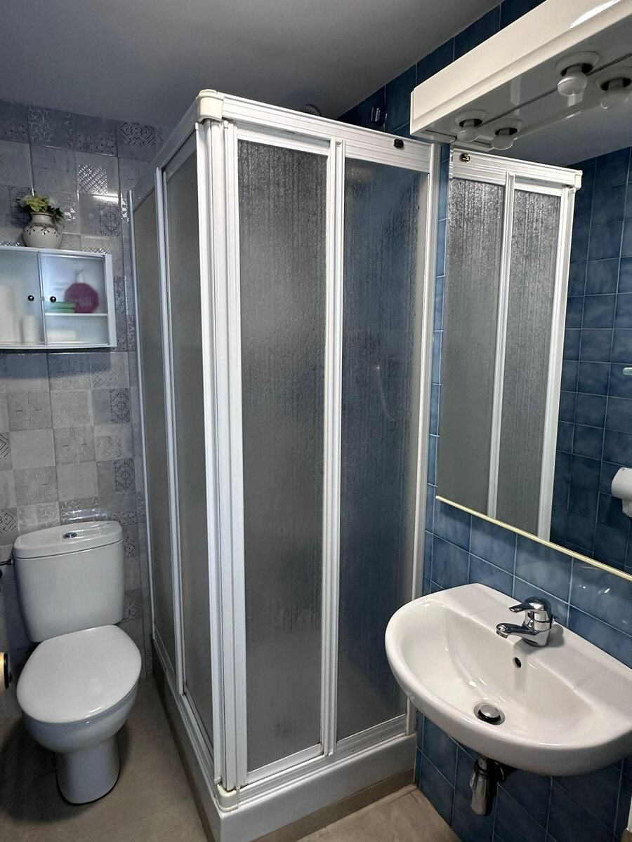 Pequeño baño con ducha de cristal esmerilado, inodoro y lavabo contra paredes de azulejos azules.