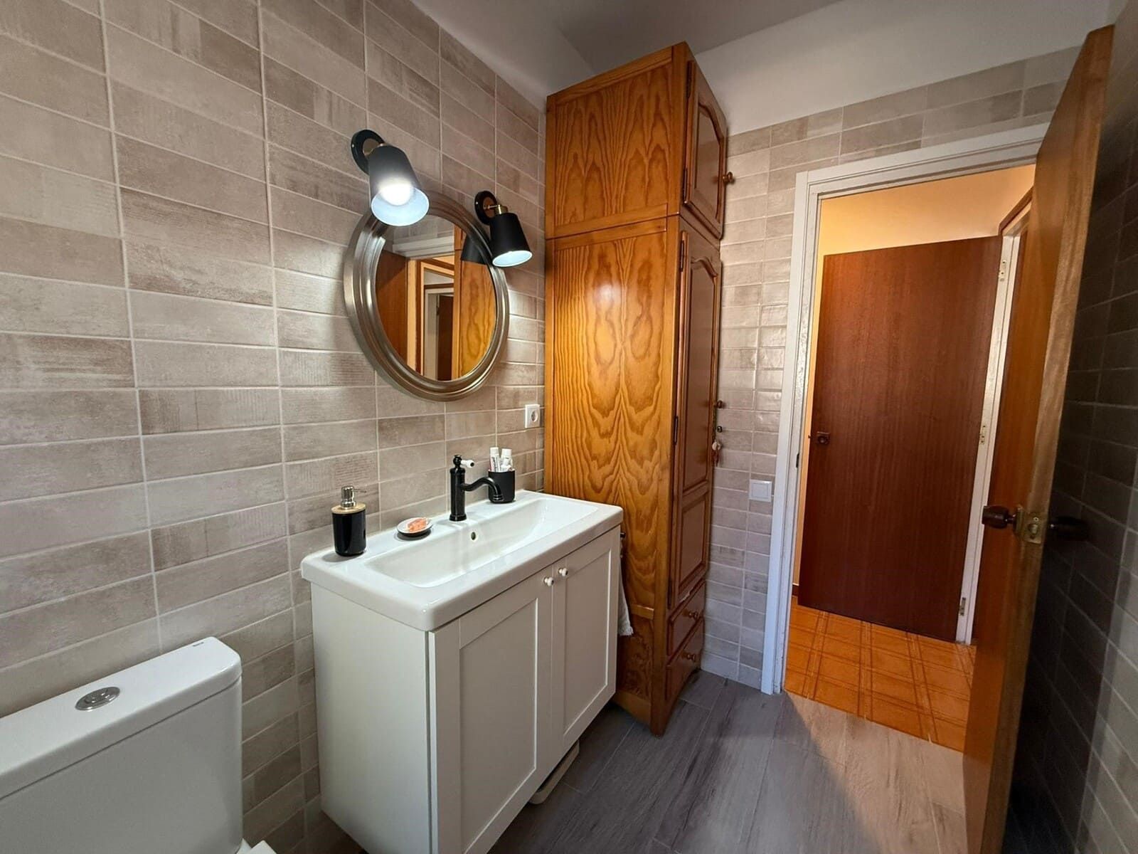 Baño con tocador blanco, mueble alto de madera y puerta abierta a otra habitación.