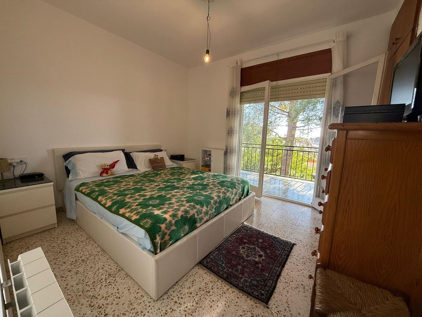 Dormitorio con cama blanca, manta estampada verde, acceso al balcón y cómoda de madera.