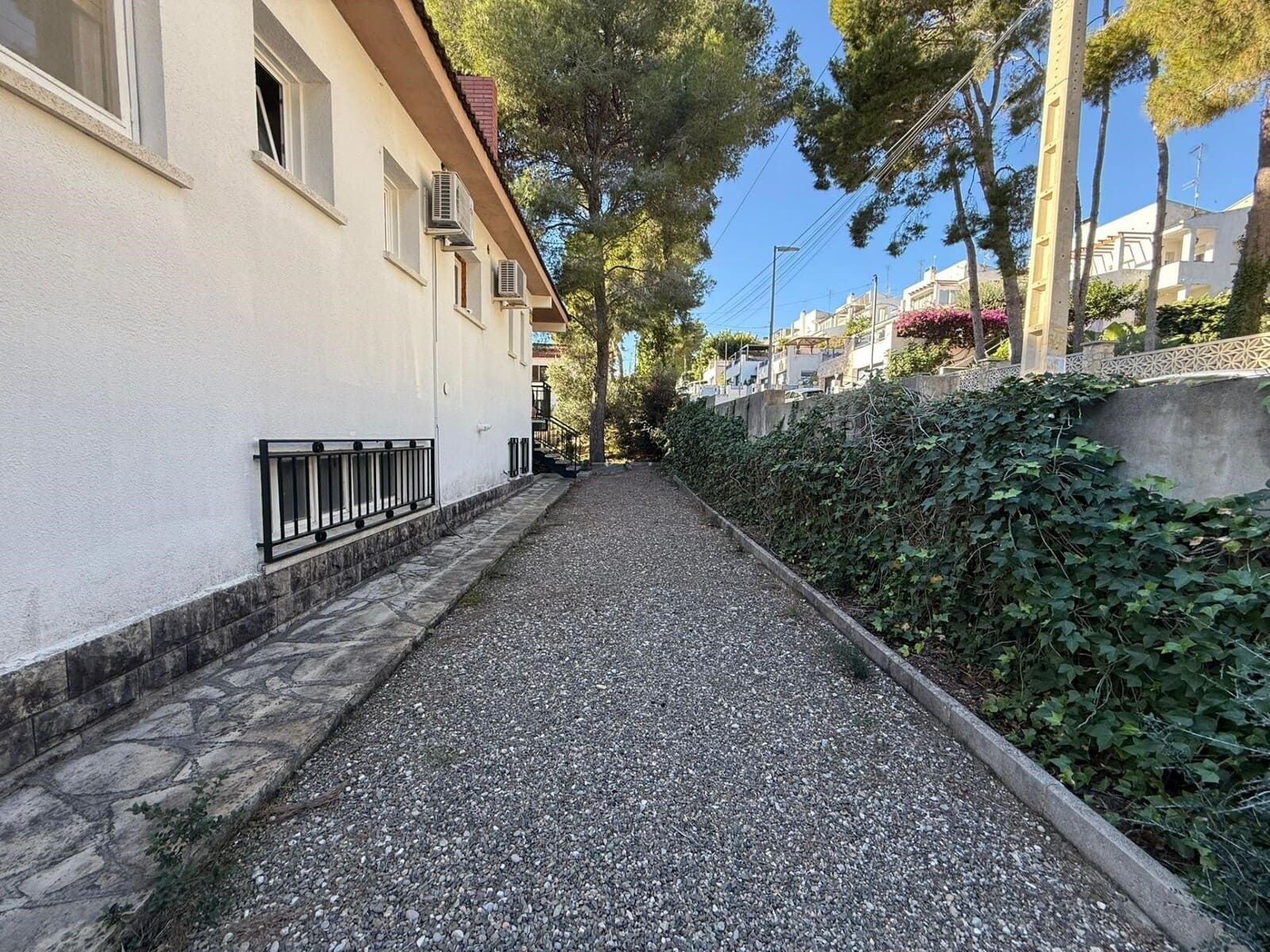Camino de grava entre un edificio blanco y una pared cubierta de hiedra verde, que conduce hacia el horizonte bajo un cielo azul.
