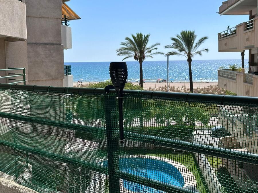Balcón con vista a la playa con palmeras y piscina, con luz solar negra fijada a la barandilla.