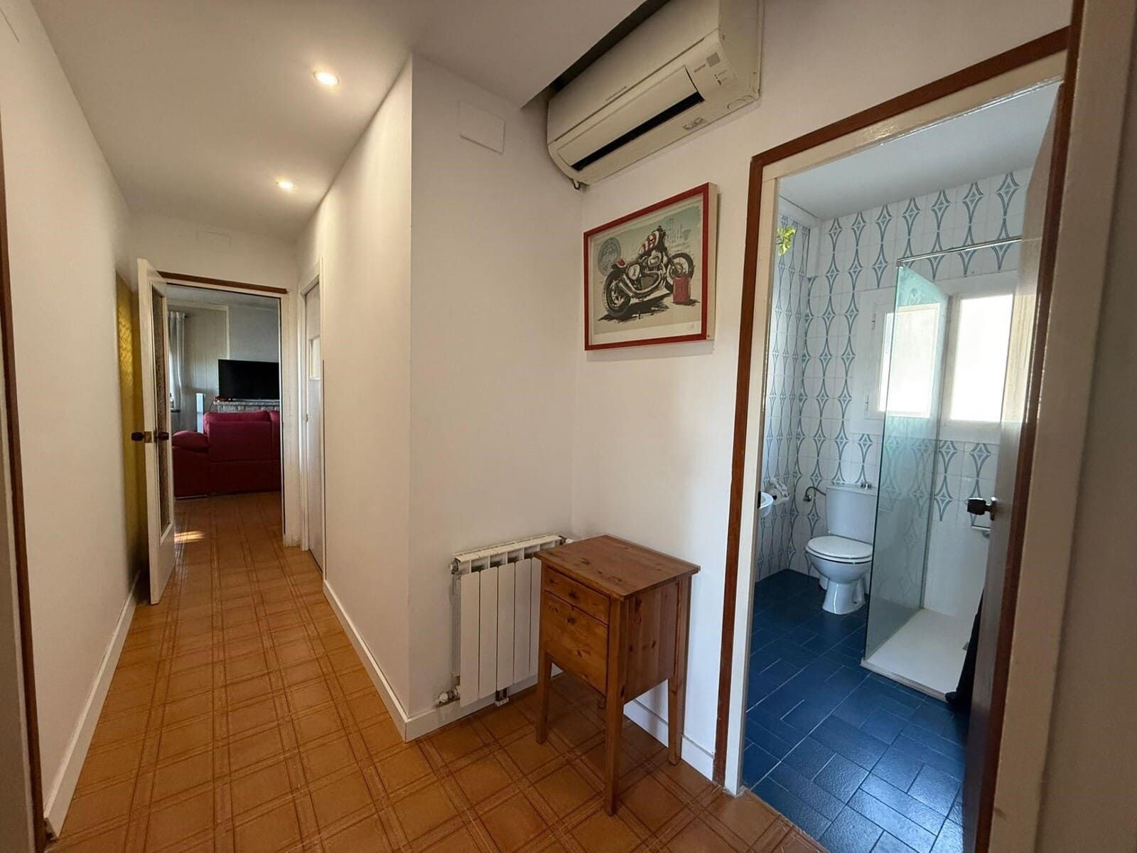 Pasillo con suelo de baldosas marrones, que conduce a una sala de estar y un baño con suelo de baldosas azules.