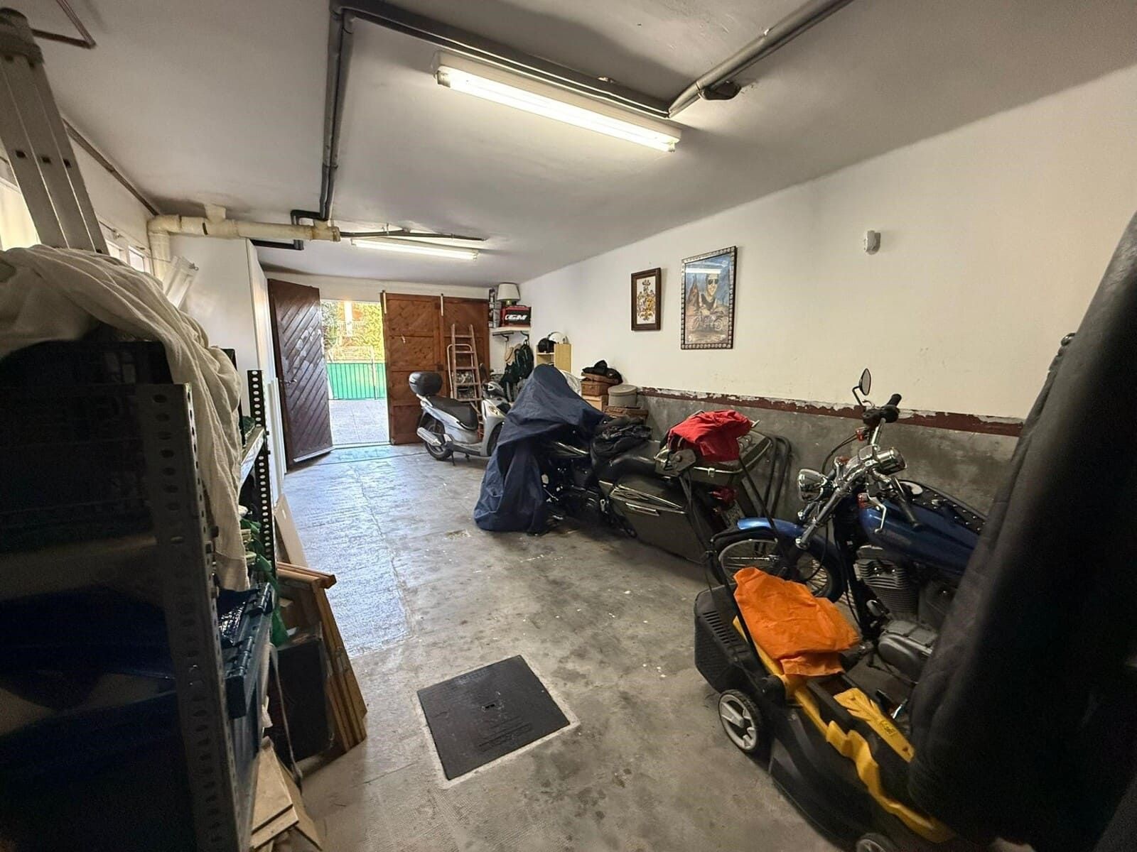 Interior de garaje desordenado. Motos, herramientas y trasteros contra las paredes. La puerta abierta revela un espacio exterior.