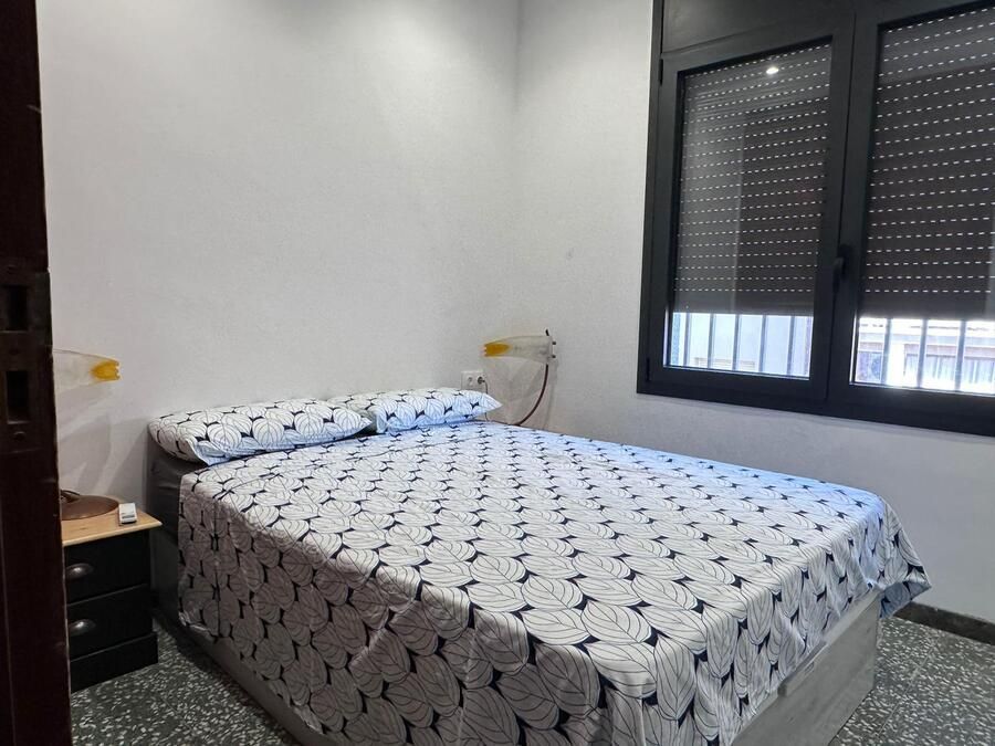 Dormitorio con cama doble, mesita de noche y ventana con persianas cerradas. Paredes blancas.