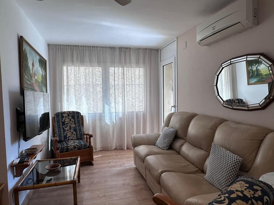 Sala de estar con sofá de cuero beige, sillón estampado, TV y ventana con cortinas transparentes.