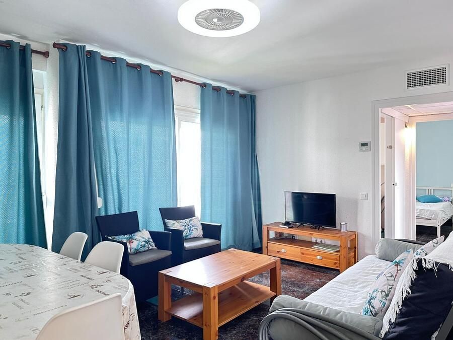 Sala de estar con cortinas azules, TV y mesa de café de madera; a través de una puerta se ve un dormitorio.