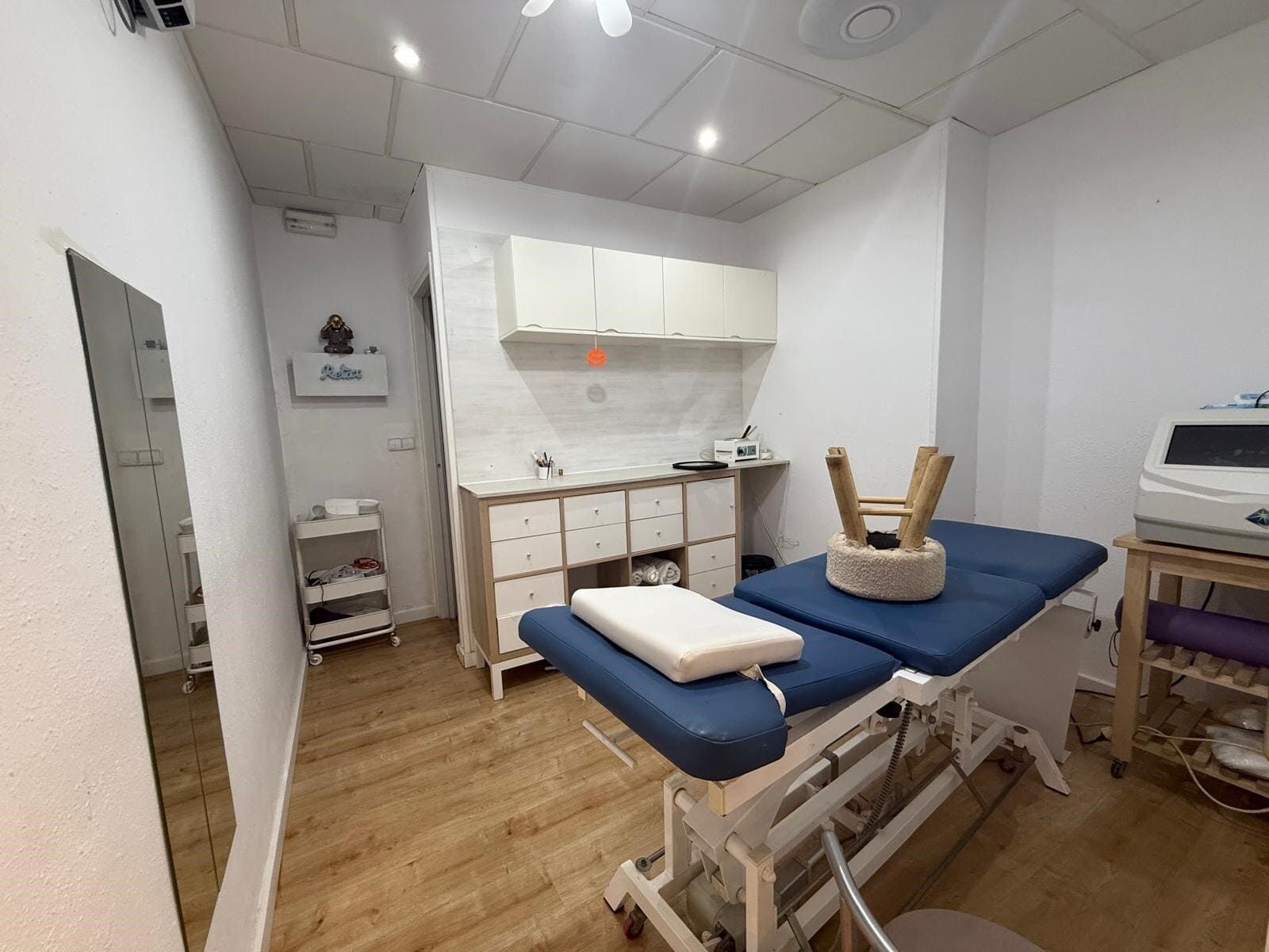 Interior de una sala de fisioterapia con una mesa de masaje azul, armarios y un espejo.