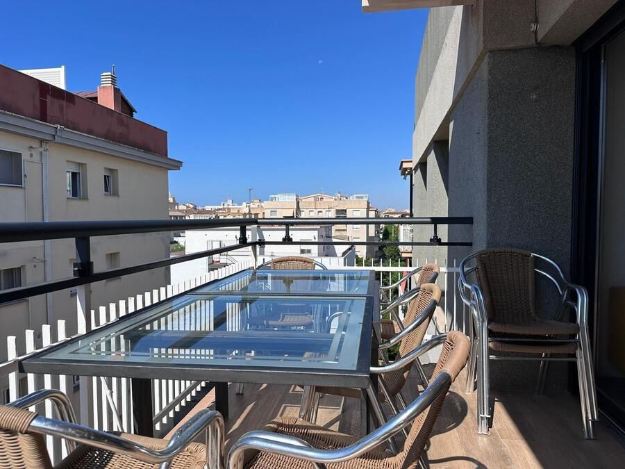 Balcón con mesa de cristal, sillas tejidas y vistas a la ciudad bajo un cielo azul.