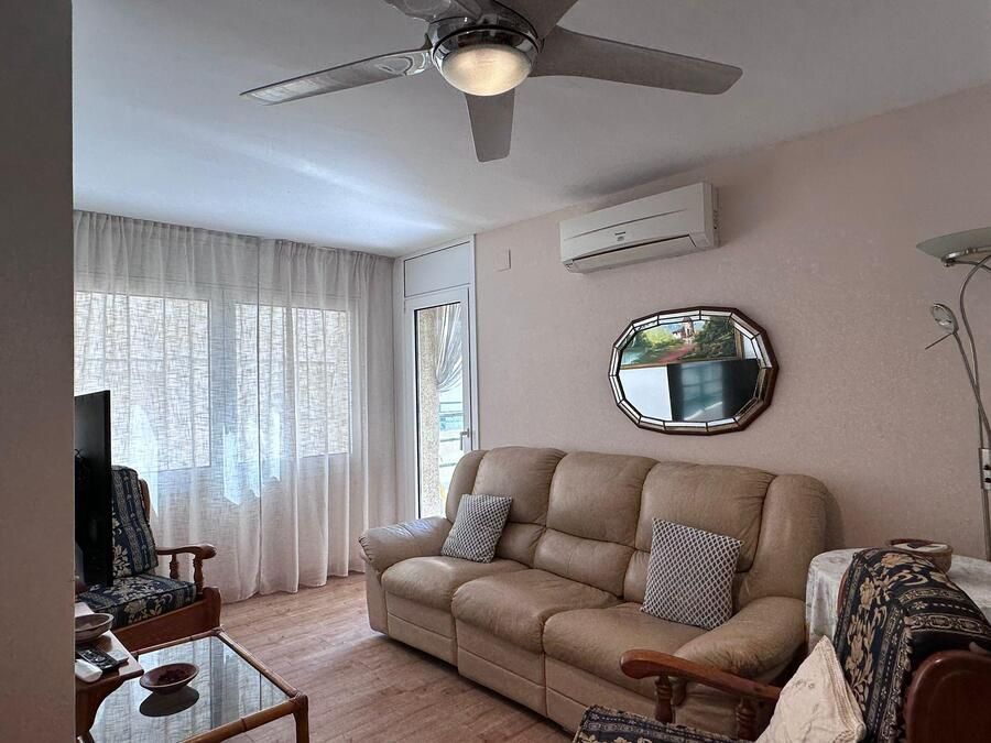 Sala de estar con sofá color canela, mesa de vidrio, ventilador de techo y ventanal con cortinas.