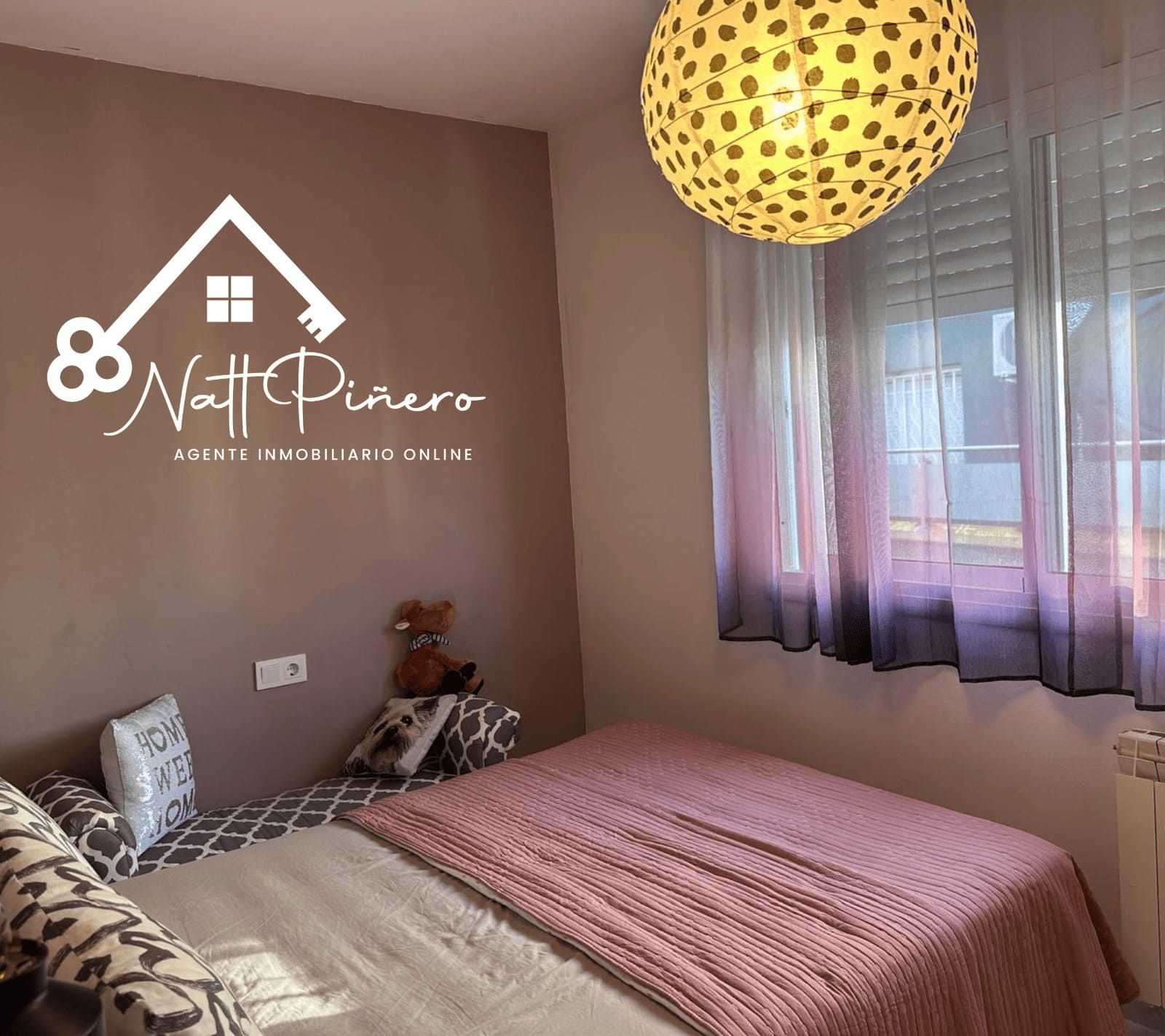 Dormitorio con cama, almohadas estampadas, una manta rosa y una pantalla de lámpara decorativa.