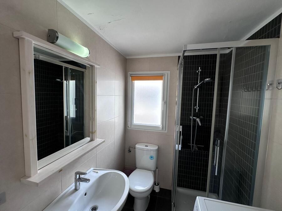 Baño con lavabo blanco, inodoro, ducha y ventana; espejo sobre el lavabo con luz fluorescente.