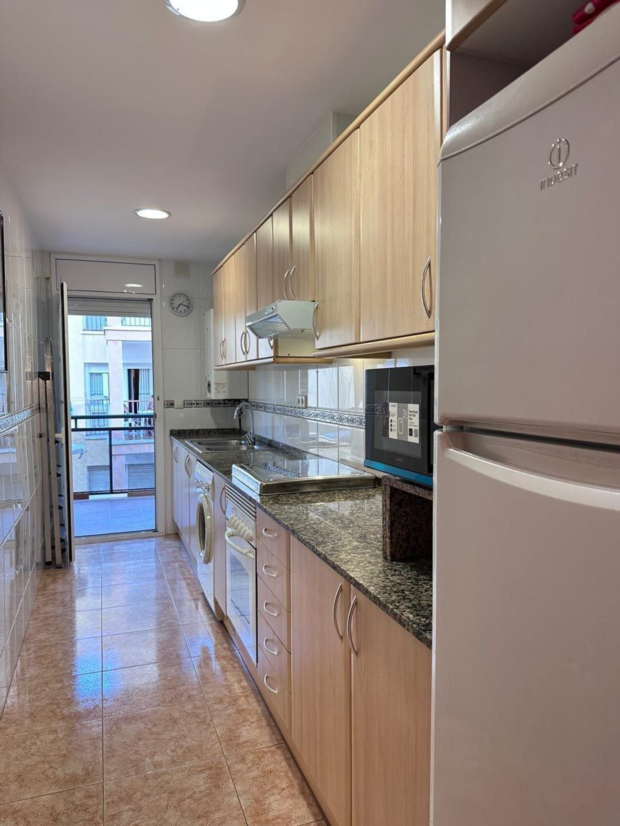 Cocina estrecha con gabinetes de madera clara, encimeras oscuras y un refrigerador a la derecha.