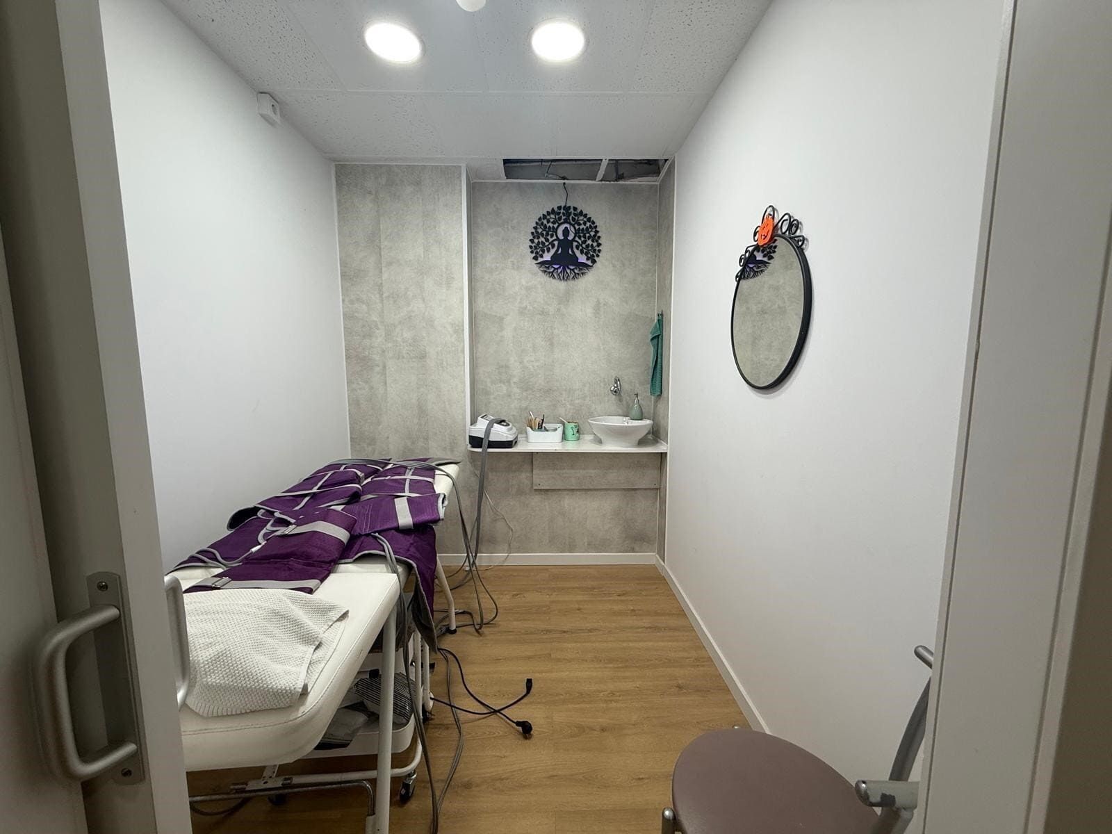 Sala de tratamientos con mesa de masajes, lavabo, espejo y reloj de pared decorativo.