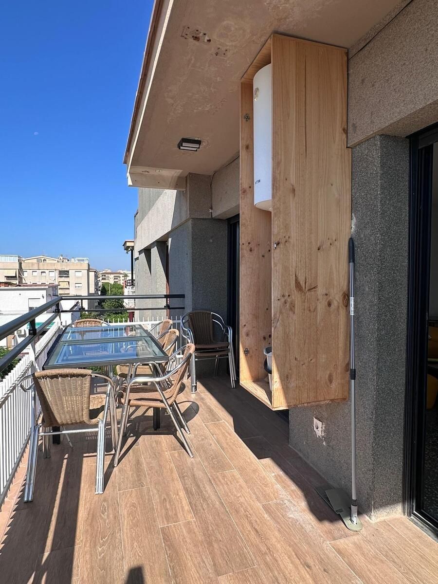 Balcón con mesa, sillas y mueble de madera adosado a la pared del edificio, bajo un cielo azul.