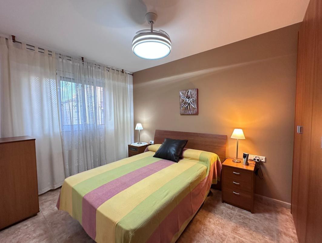 Dormitorio con cama, mesitas de noche, armario, ventana con cortinas transparentes y ventilador de techo.