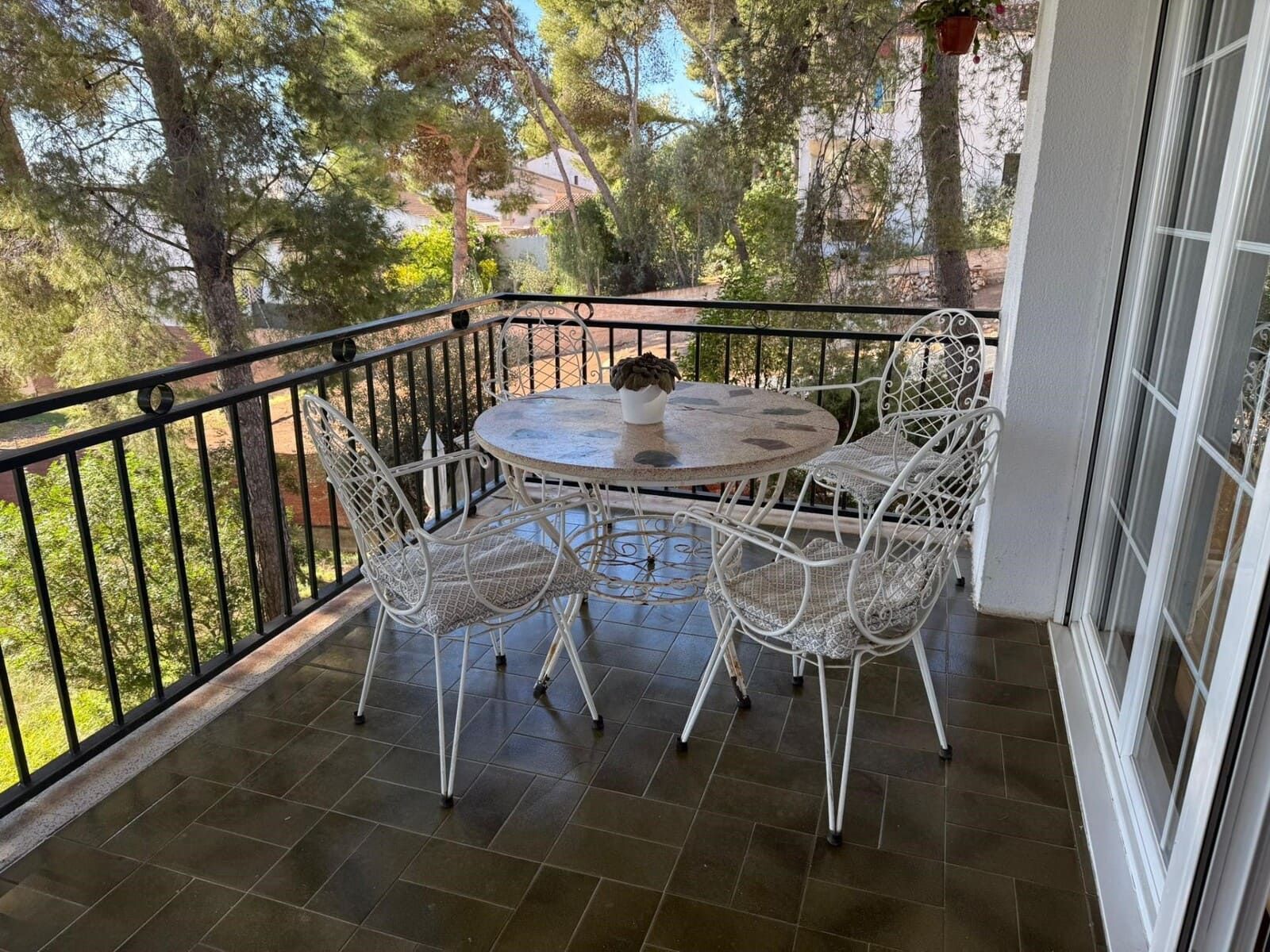 Balcón con sillas de alambre blanco, mesa redonda, barandilla negra y árboles.