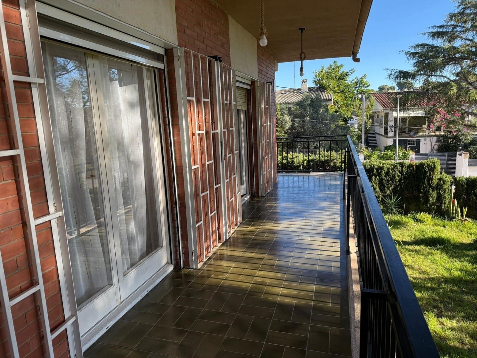 Balcón de una casa de ladrillo con piso de baldosas oscuras, puertas de vidrio y barandilla de metal negro.