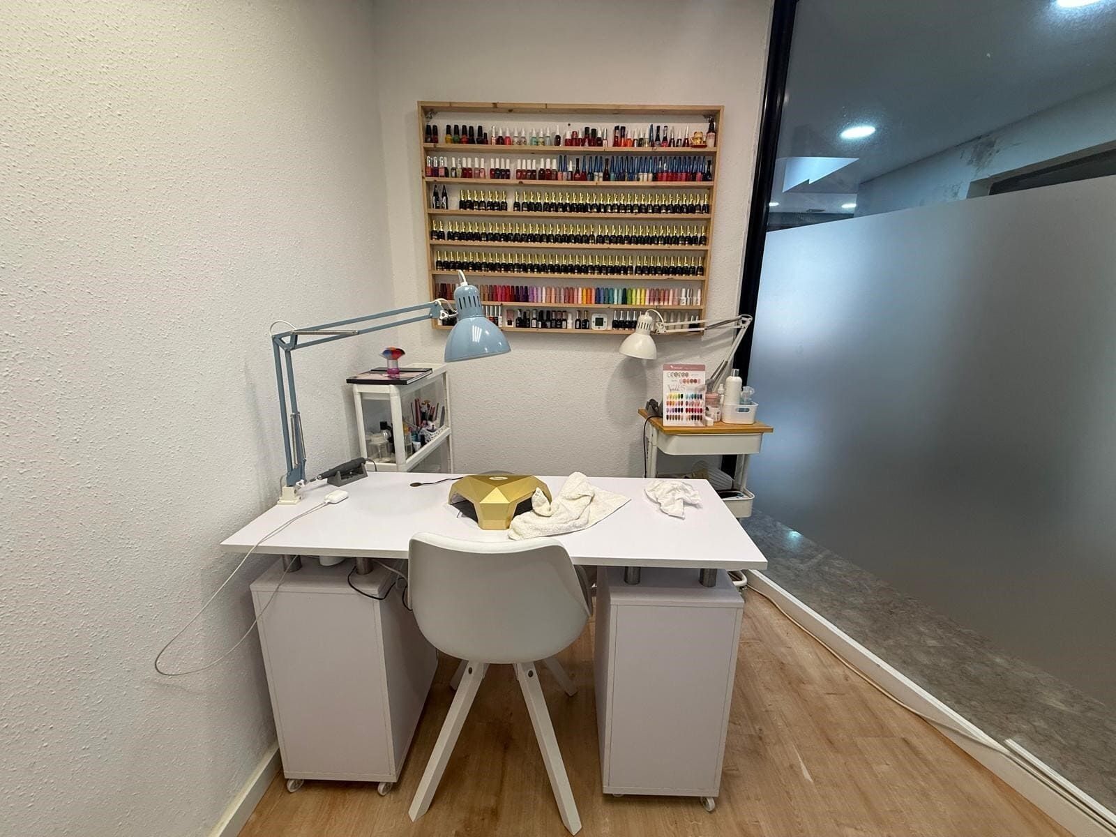 Salón de uñas con escritorio blanco, silla y estantes con esmaltes de uñas. Una lámpara sobre el escritorio.