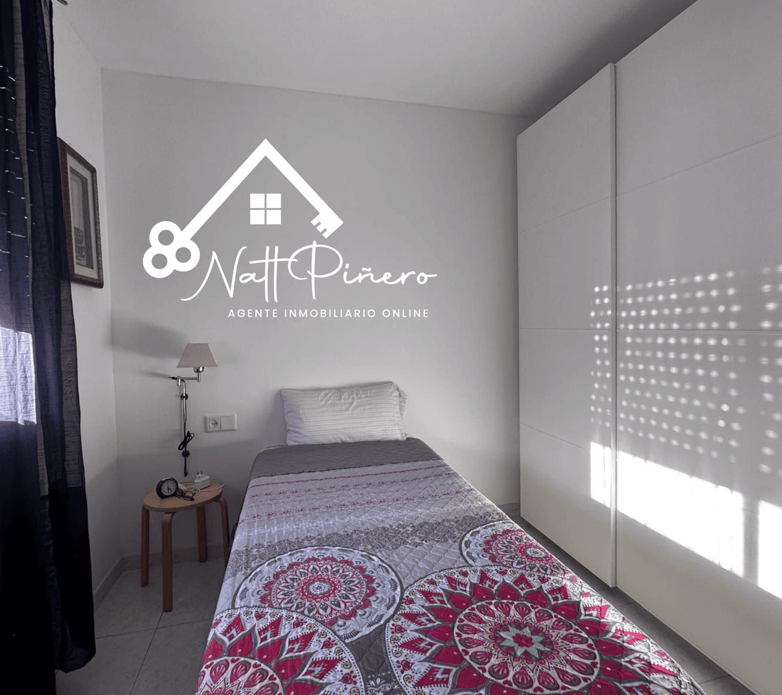 Pequeño dormitorio con una cama individual, armario blanco, pequeño taburete de madera y una lámpara de mesa.