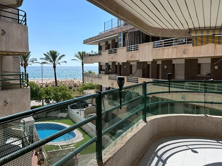 Vista desde el balcón: playa, piscina, edificios y palmeras bajo un cielo azul soleado.