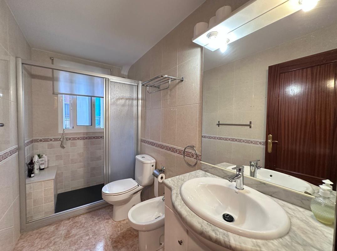 El baño cuenta con lavabo, inodoro y bidé blancos. Se ve una puerta de ducha de cristal. La habitación es beige y marrón.