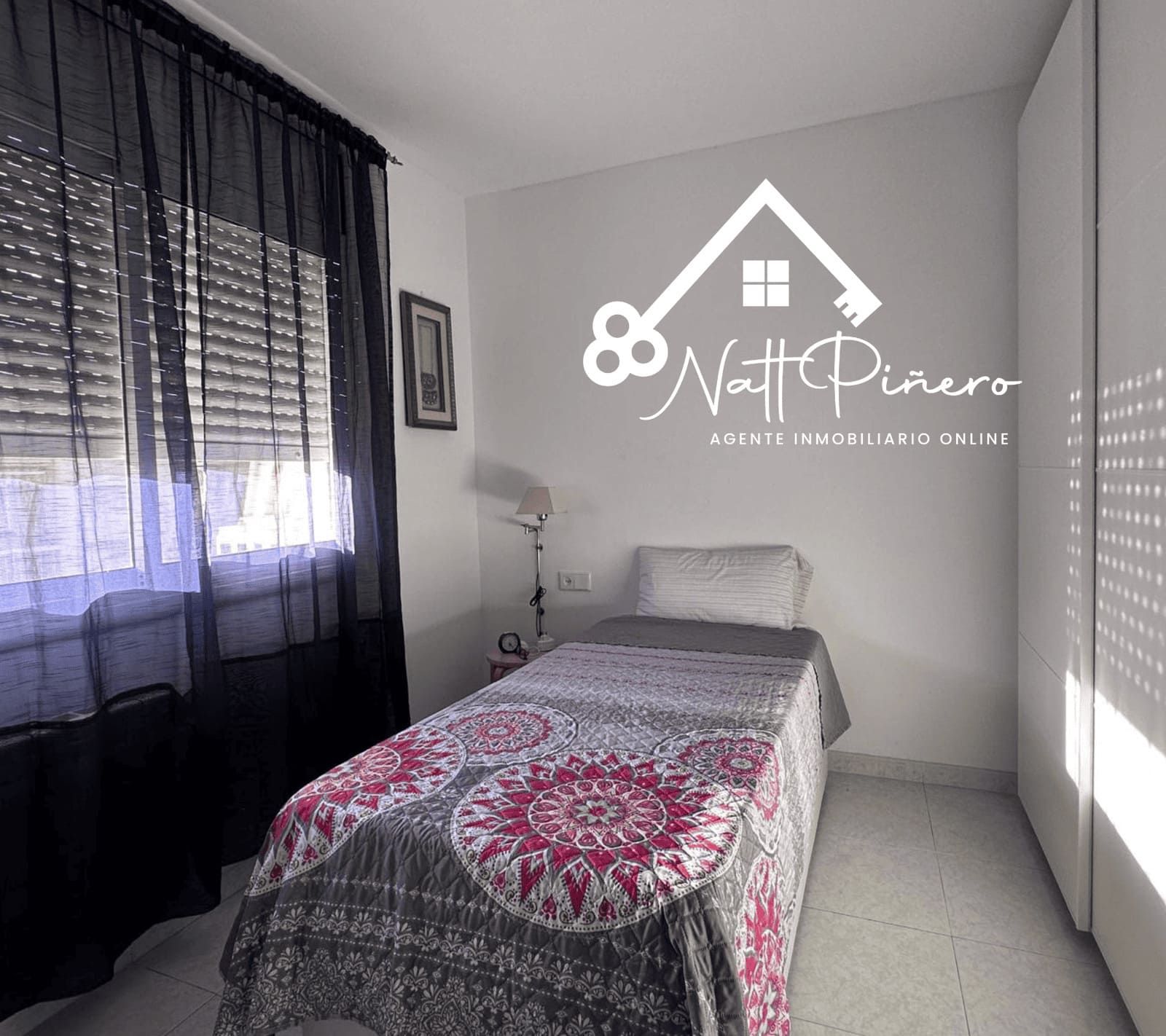 Dormitorio con cama individual, ventana con cortina oscura y armario blanco. Hay un logotipo en la pared.